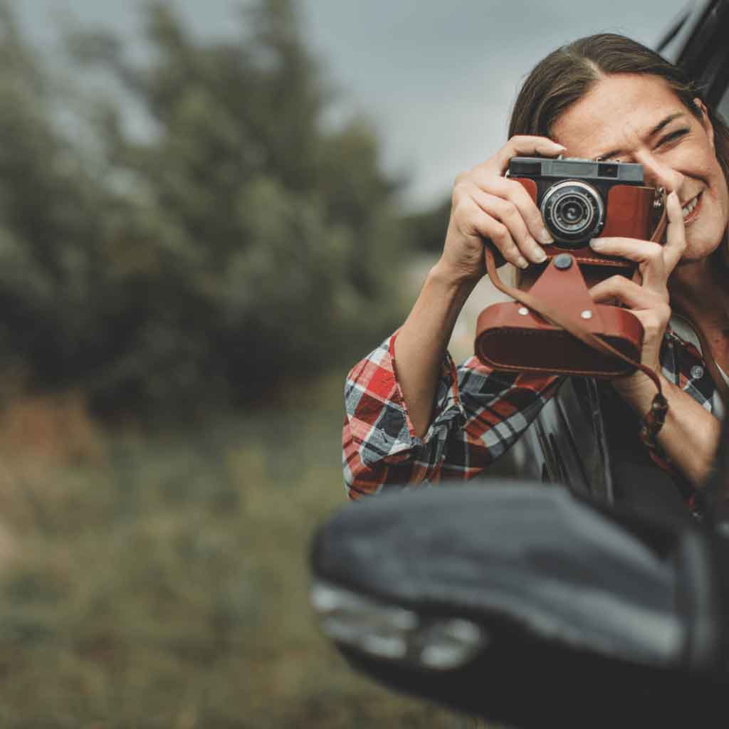 A cheerful woman leaning out of a car window, snapping a photo with a retro-style camera